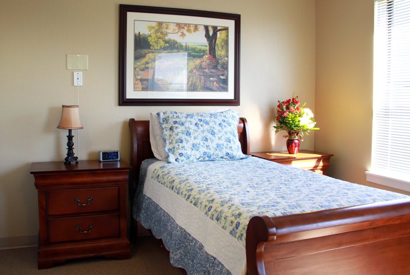 A cozy bedroom with a single bed featuring floral bedding, a nightstand with a lamp, and a framed painting above the bed.