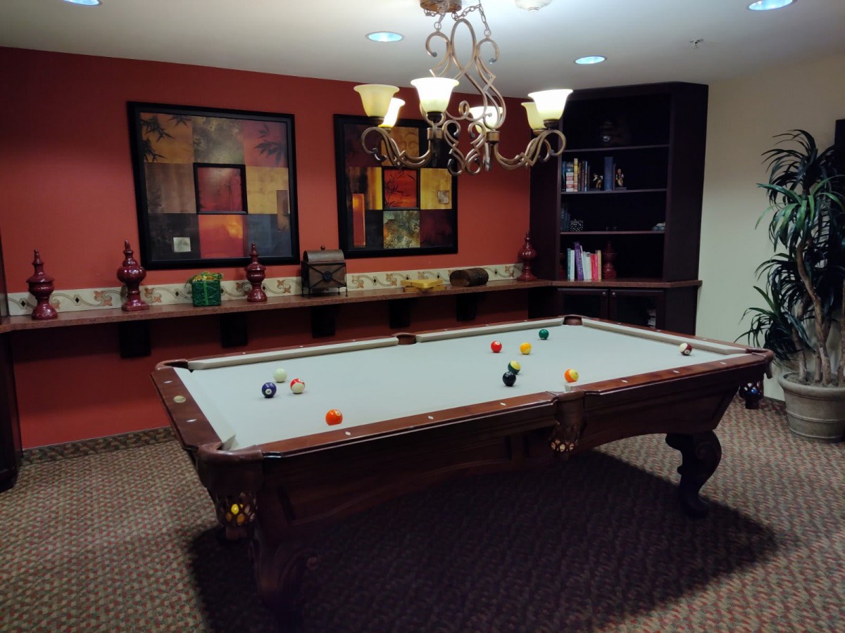 A cozy game room featuring a pool table under a chandelier, with artwork and a bookshelf in the background.