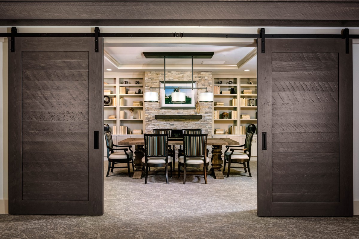 A modern conference room with sliding doors, featuring a central table surrounded by chairs, bookshelves along the walls, and a TV screen mounted above.