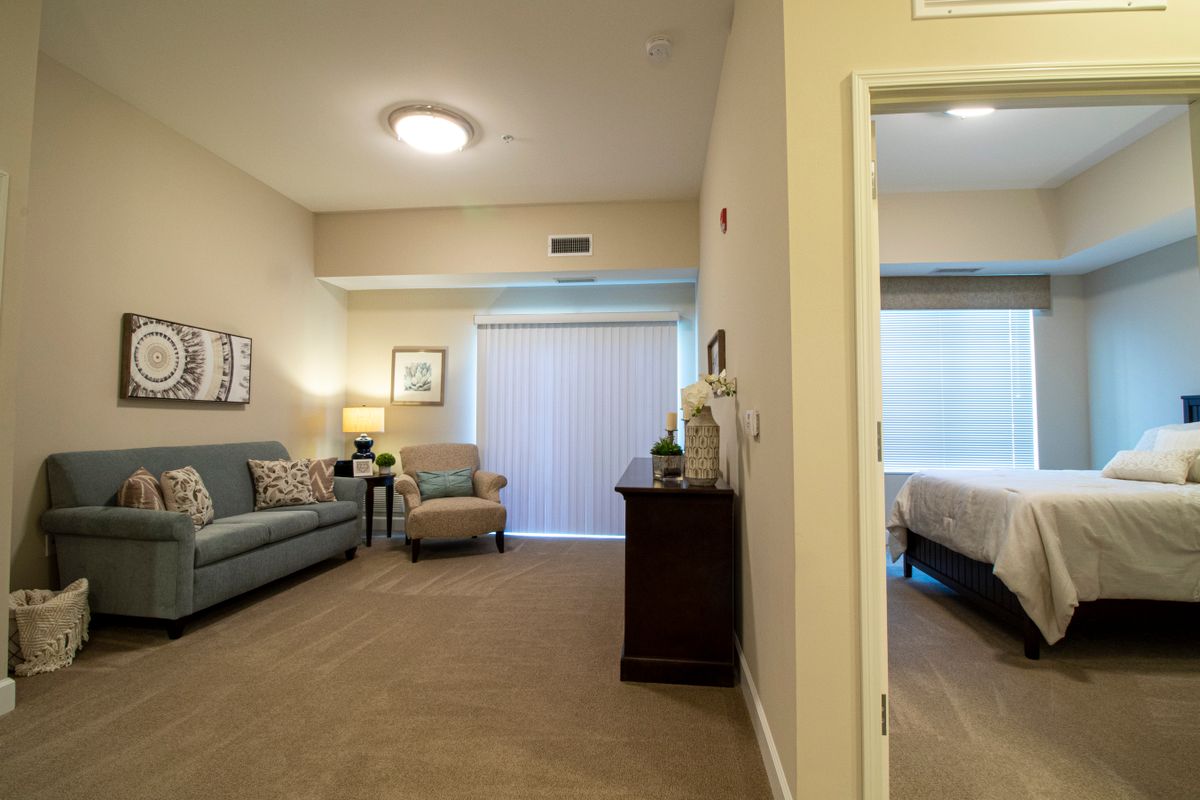 A cozy apartment interior featuring a living room with a sofa and lamp, leading into an adjoining bedroom.