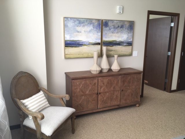 A cozy room corner with a wooden cabinet, decorative vases, and landscape artworks above, accompanied by a cushioned chair.
