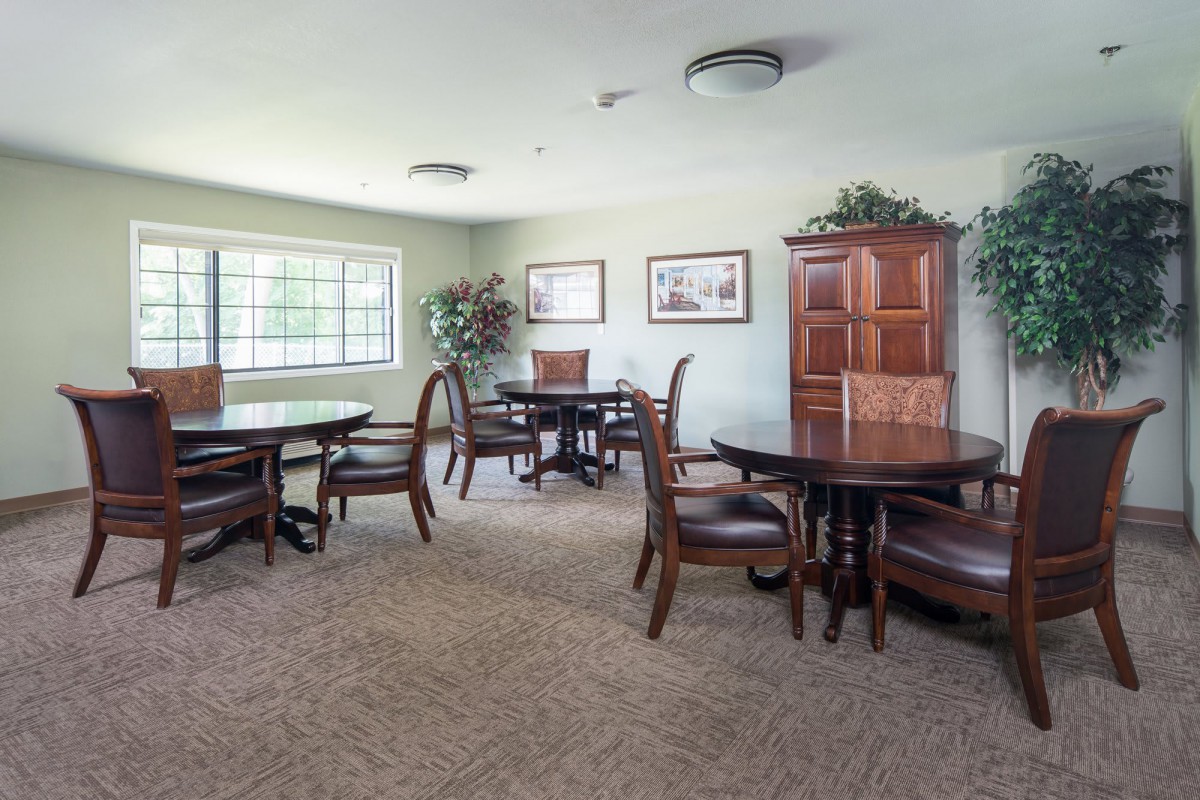 A spacious room with round wooden tables, chairs, and a large window allowing natural light to fill the space.