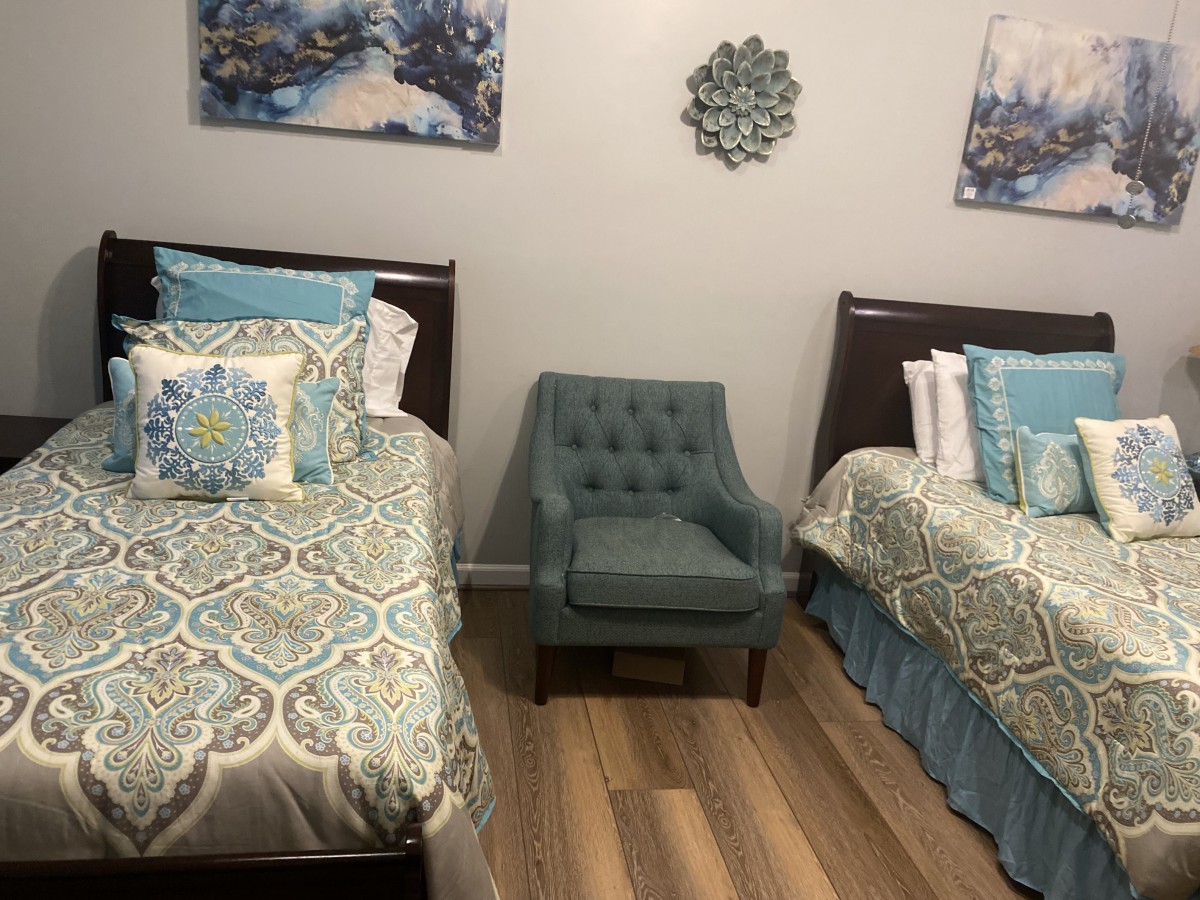 A cozy bedroom with two twin beds and a teal armchair, featuring patterned bedding and wall art.