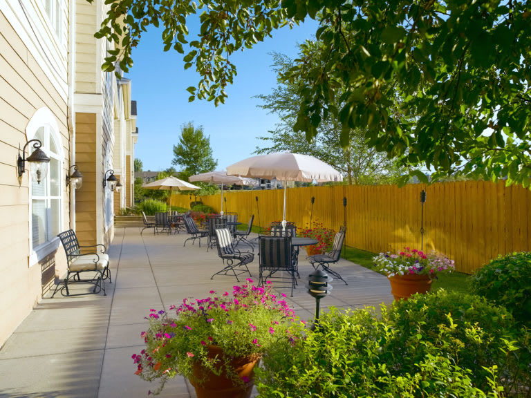 A sunny patio with outdoor seating, tables, and umbrellas surrounded by a garden and a wooden fence.