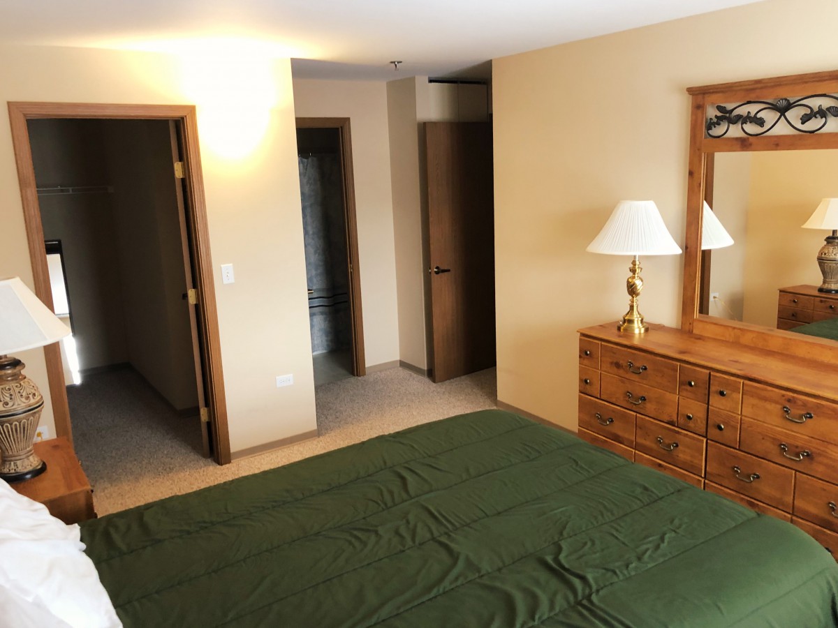 A cozy bedroom featuring a bed with a green blanket, wooden dresser with a mirror, and a lamp.