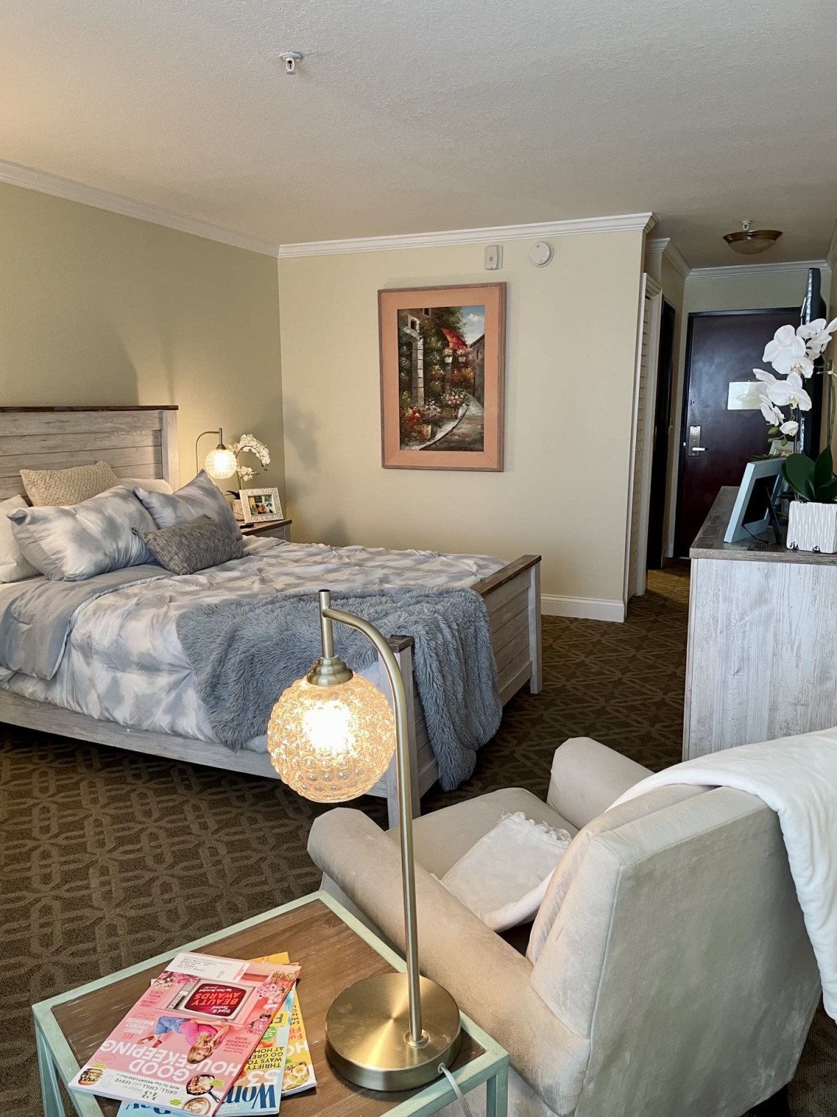 A cozy hotel room with a comfortable bed, nightstand, lamp, and decorative artwork on the wall.