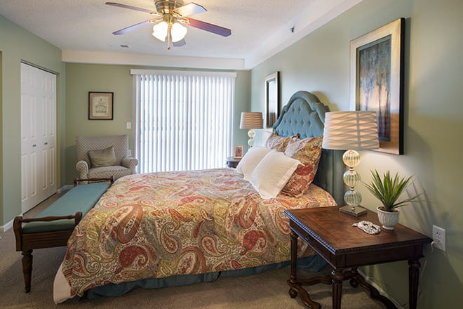 A cozy and well-decorated bedroom featuring a large bed with colorful bedding, a nightstand with a lamp, and a comfortable chair. A ceiling fan hangs overhead, and sunlight filters through vertical blinds.