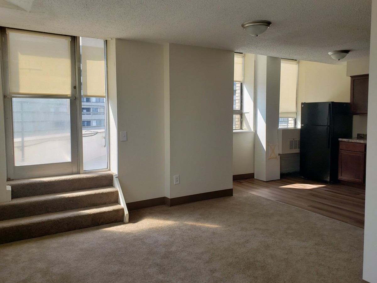An empty apartment interior with carpet flooring, natural light streaming through large windows, and a small set of stairs leading to a balcony.