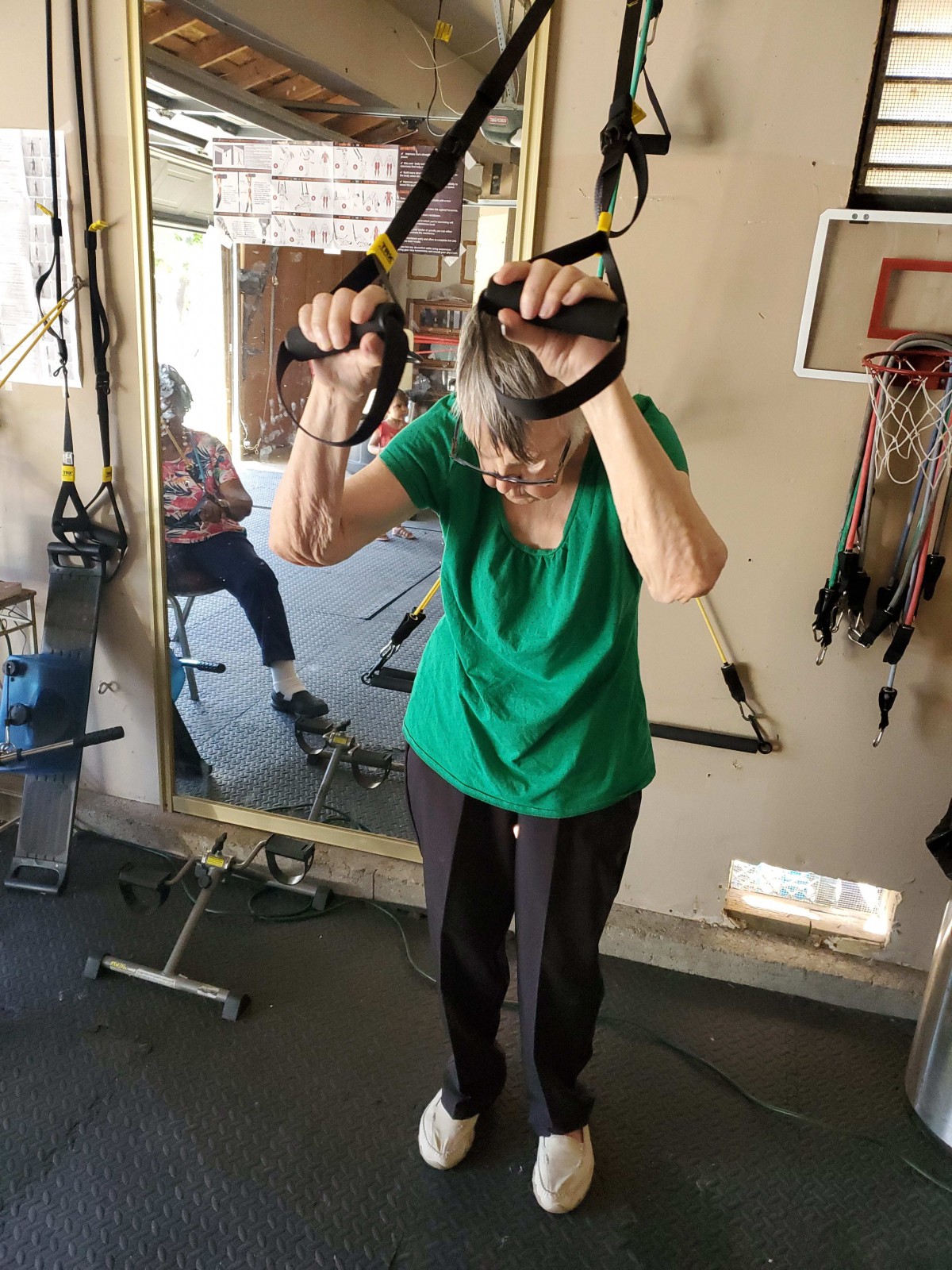 An elderly woman exercising with TRX straps in a home gym setting.