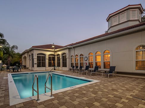 An outdoor swimming pool with a building in the background, illuminated by evening lights.