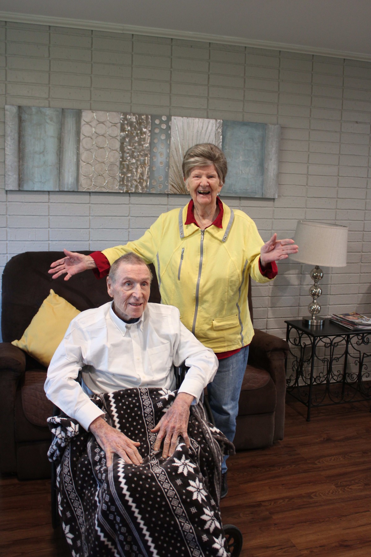 An elderly couple in a living room, with one person sitting in a wheelchair and the other standing beside them, both smiling.