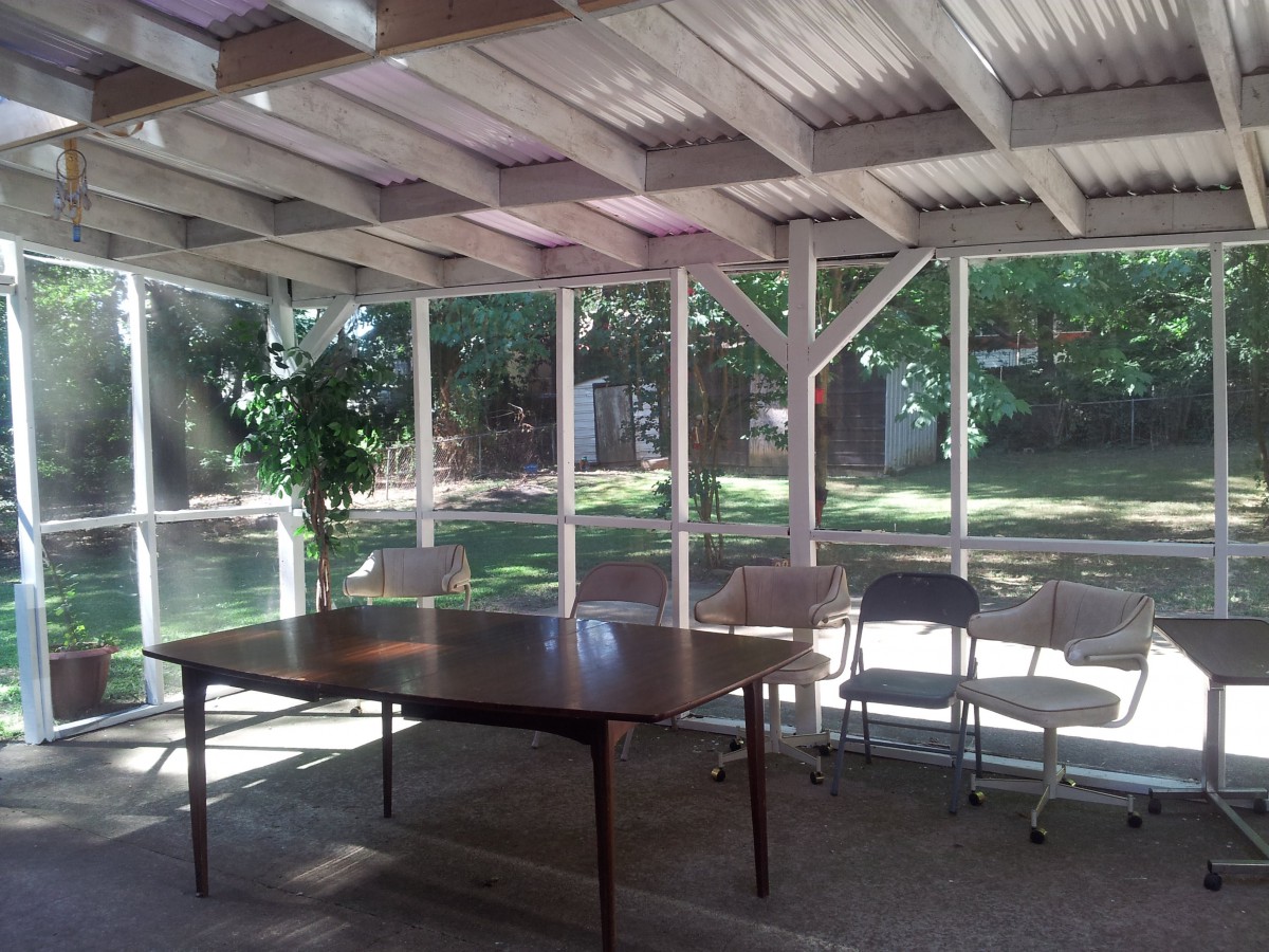 A sunlit room with a table and chairs, surrounded by large windows and greenery.