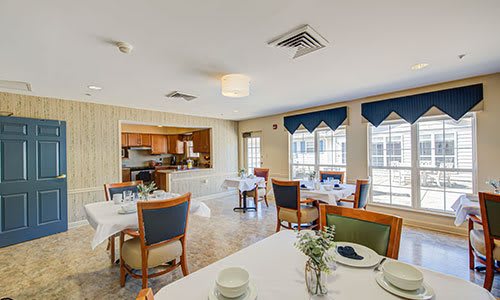 A well-lit dining area with neatly arranged tables and chairs, adjacent to a kitchen space.