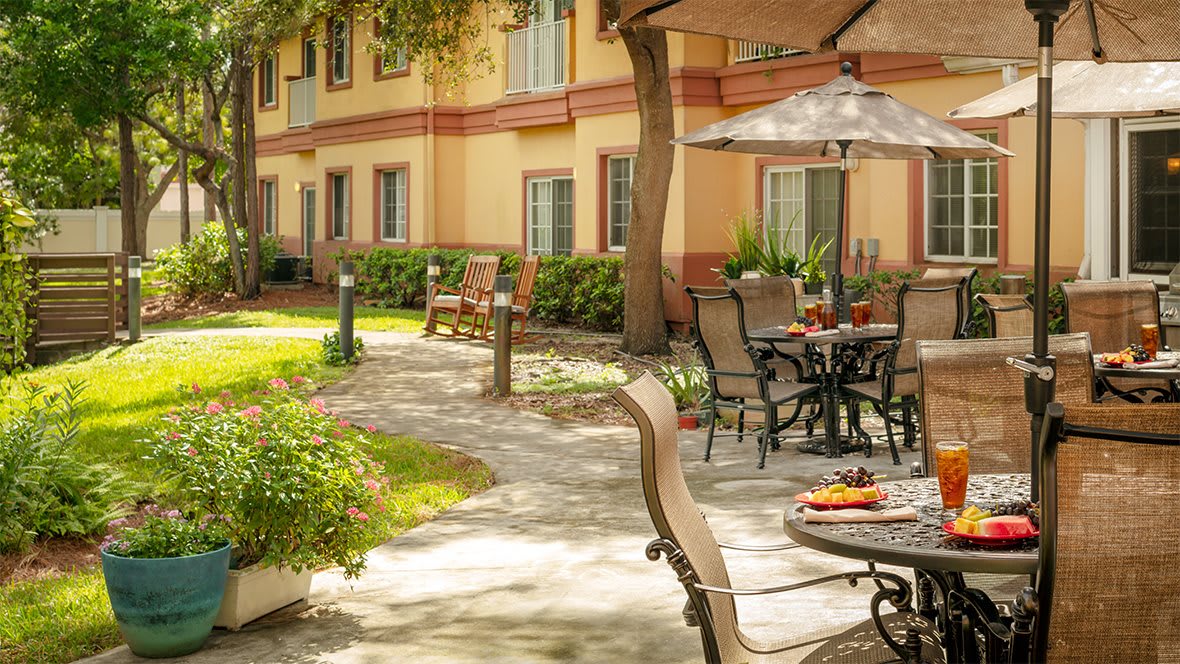 A peaceful outdoor patio area with tables, chairs, and umbrellas on a stone pathway, surrounded by greenery and adjacent to a building.