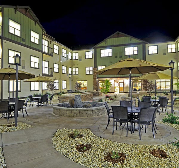 A well-lit courtyard at night featuring tables, chairs, and umbrellas in front of a multi-story building.