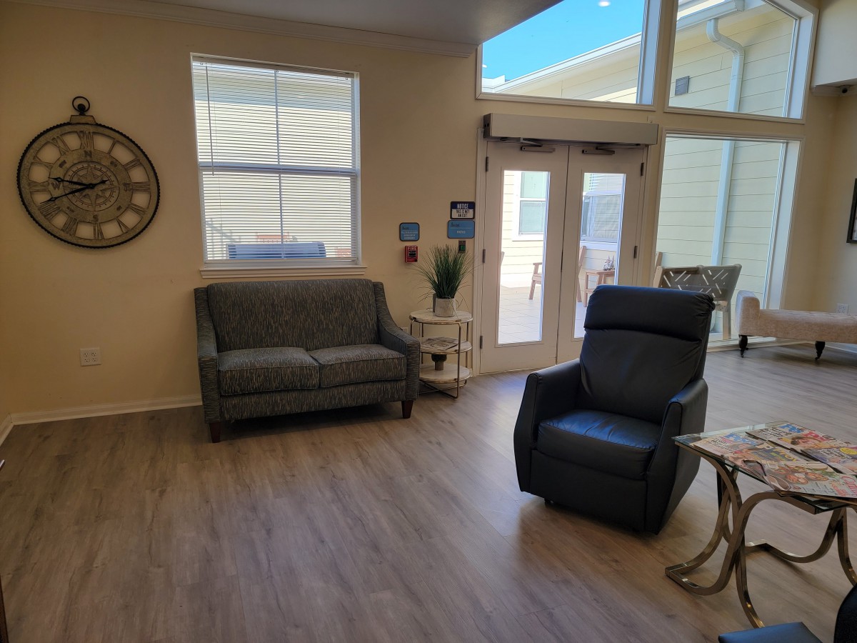 A cozy living room with a sofa and a recliner chair, featuring natural light from large windows.