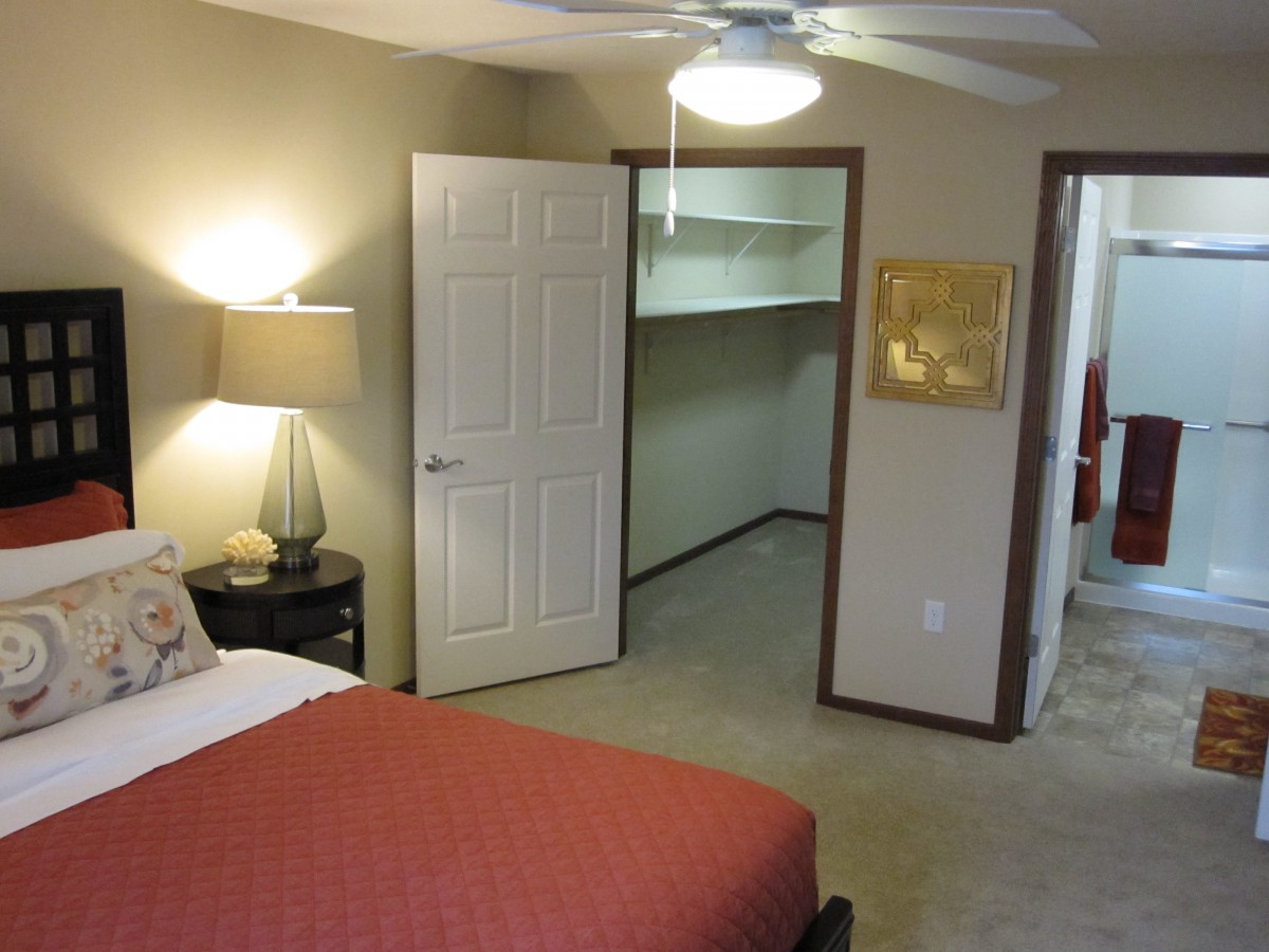 A cozy bedroom featuring a bed with red bedding, a bedside lamp, and an open closet door revealing shelving space.