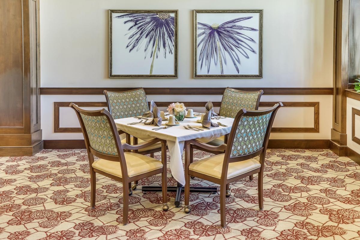 An elegantly set dining table with four chairs, complemented by artwork on the wall and patterned carpeting.