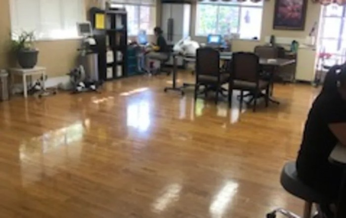 A spacious office with desks, chairs, and computers, featuring wooden flooring and large windows.