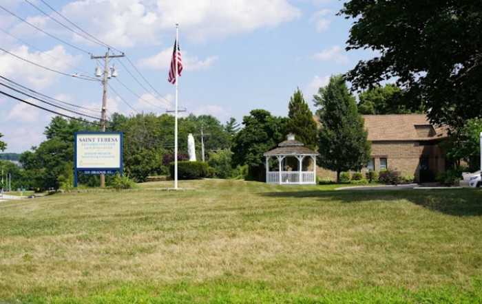 A grassy area featuring an American flag, a gazebo, and a building under a partly cloudy sky.