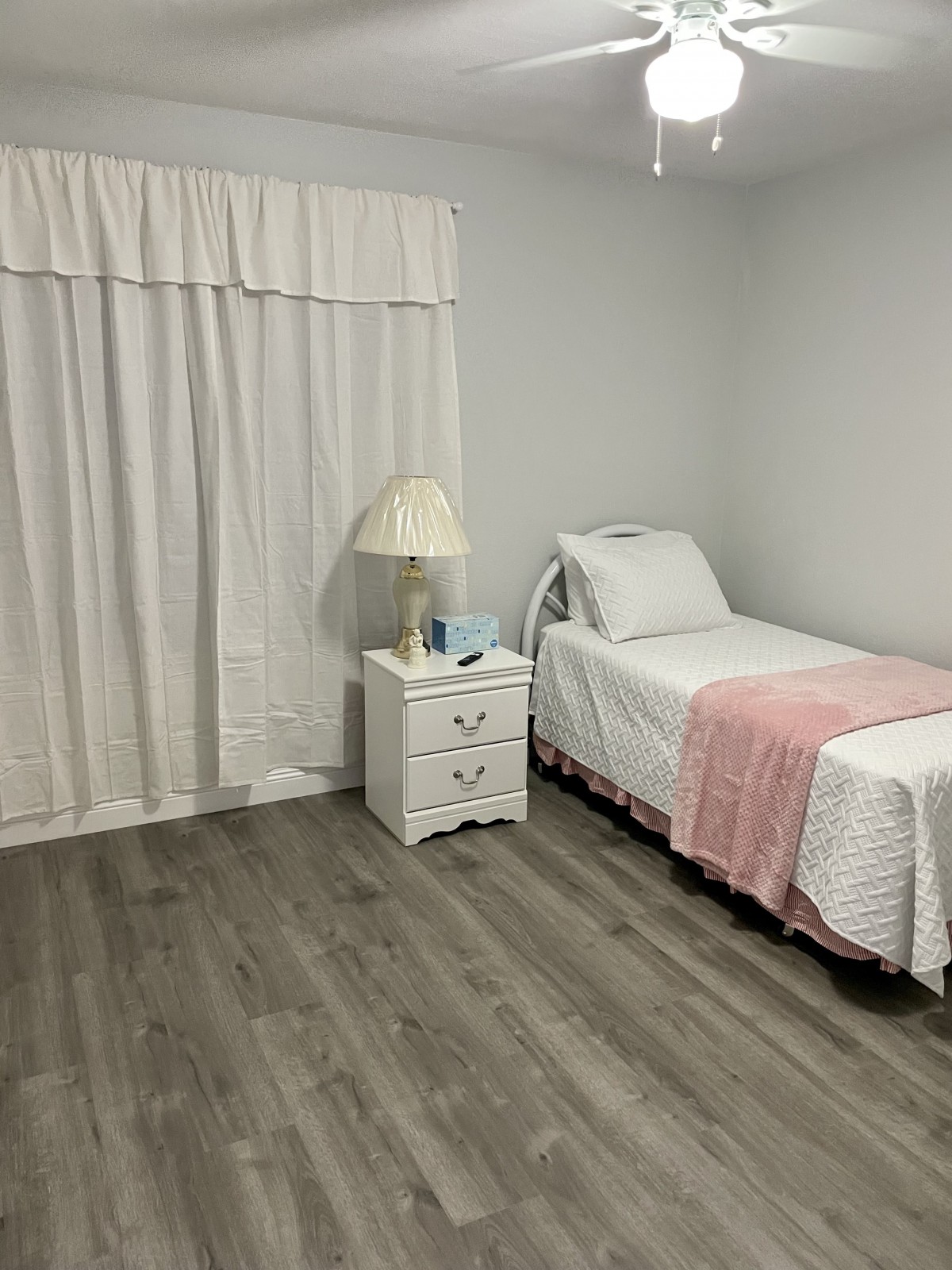 A minimalist bedroom with a single bed, pink blanket, and white curtains.