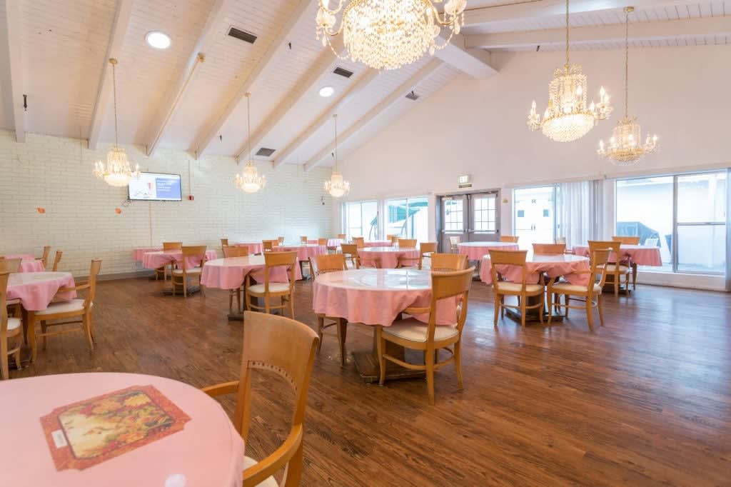 A spacious dining area with pink tablecloth-covered tables, wooden chairs, and elegant chandeliers.