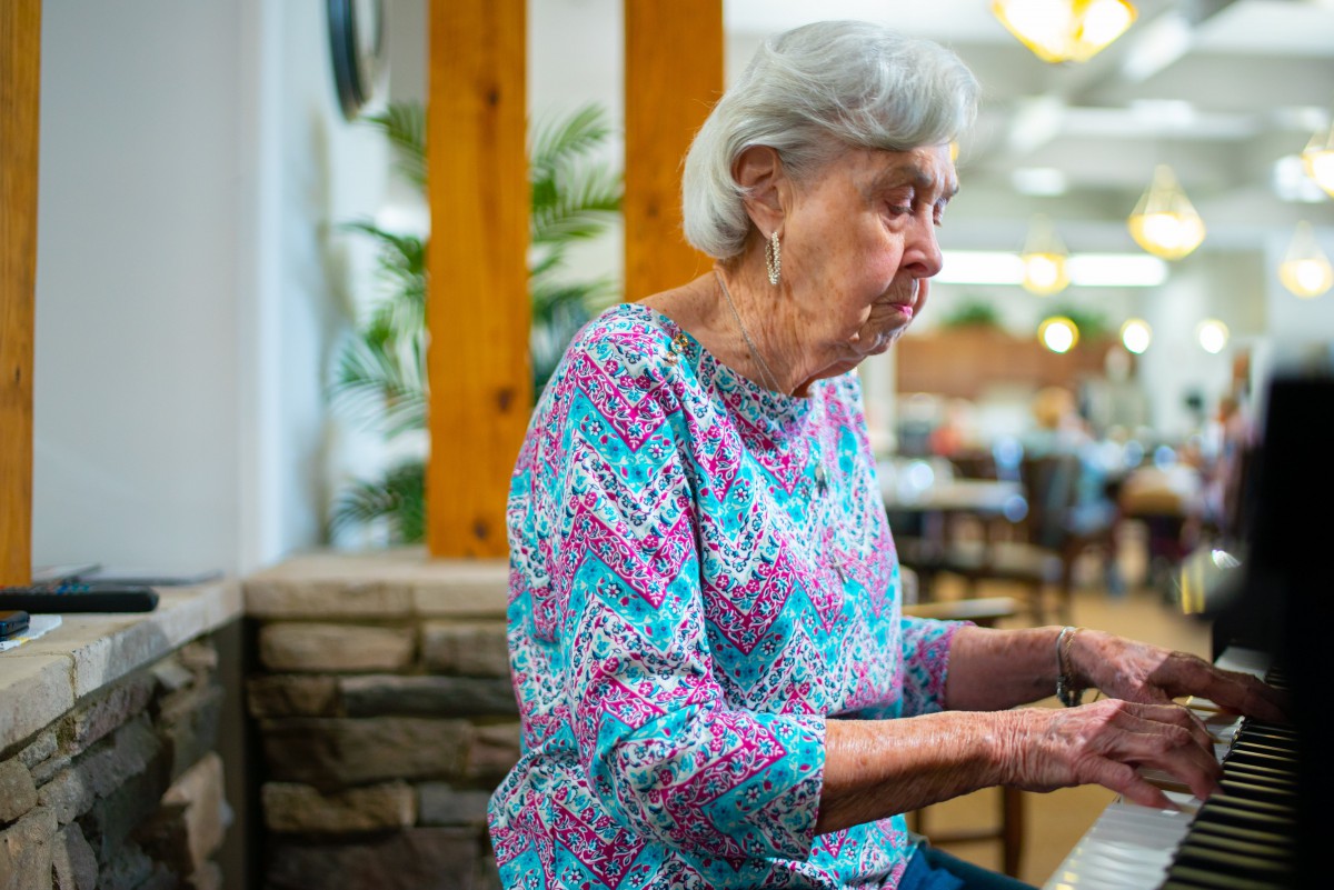An elderly woman is playing the piano indoors, wearing a colorful shirt.