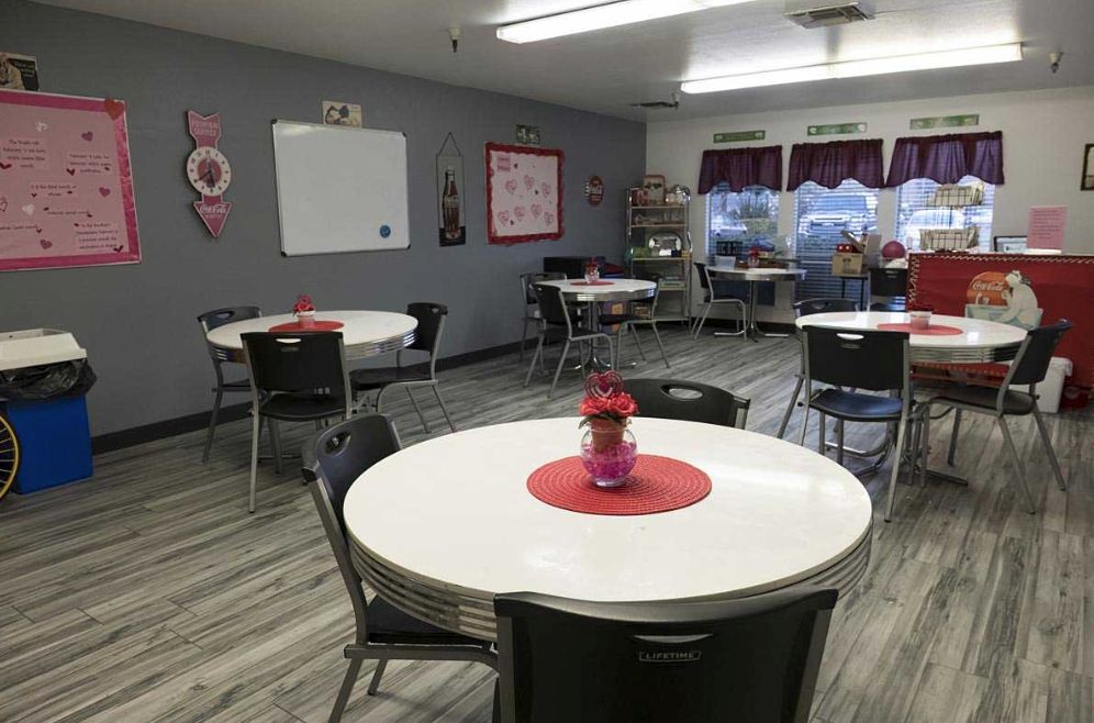 A well-organized classroom with round tables and chairs, featuring a whiteboard on the wall and windows allowing natural light.