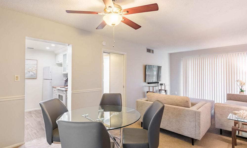 A modern apartment interior featuring an open-plan dining and living area with a ceiling fan, glass dining table, upholstered chairs, comfortable sofa, and mounted television.