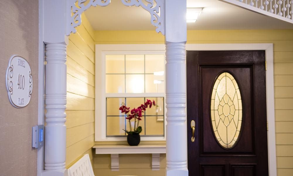 A cozy front porch with a yellow exterior and decorative trim features a dark wooden door and a window with a blooming orchid on the sill.