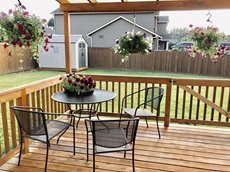 A cozy backyard patio with a wooden deck, complete with a table and chairs surrounded by colorful flowers and hanging plants.