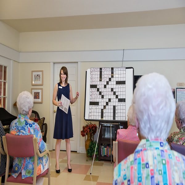 A woman is leading a crossword puzzle activity with a group of elderly participants in a community room.