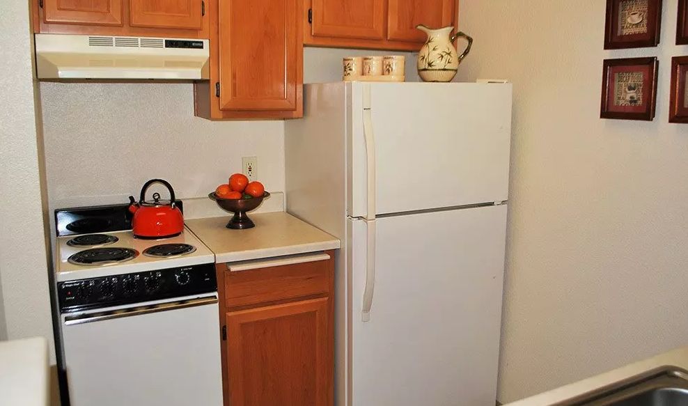 A compact kitchen space featuring a white refrigerator, wooden cabinets, and a countertop with a fruit bowl and teapot.