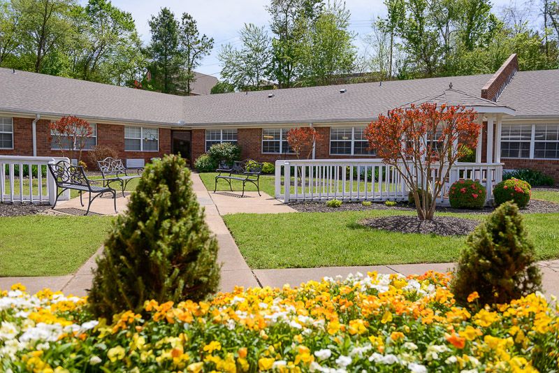 A beautiful garden with vibrant flowers in front of a brick building, surrounded by trees and benches.
