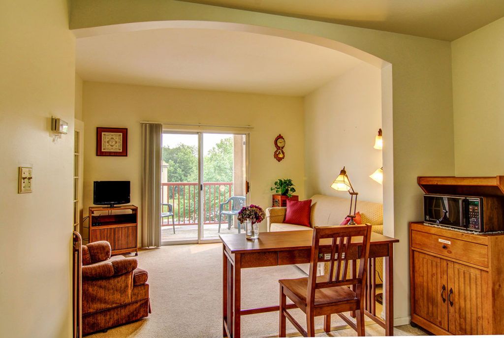 A cozy living room with a desk, comfortable seating, and natural light from the balcony door.