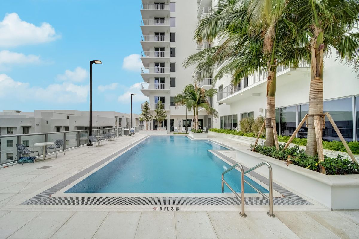 An outdoor swimming pool beside a modern apartment building, surrounded by palm trees.