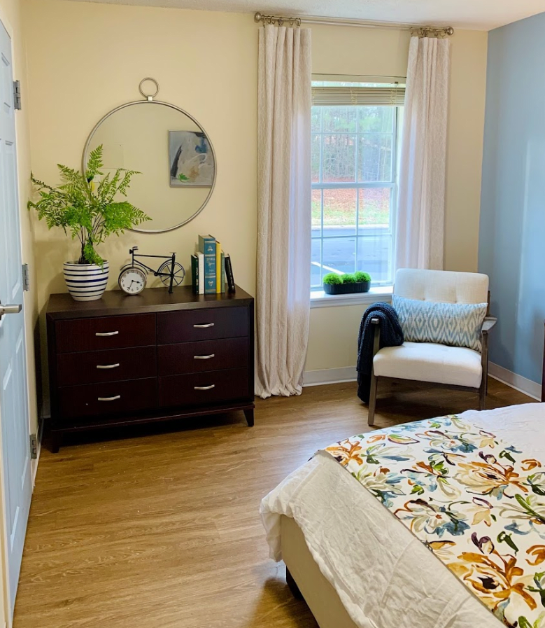 A cozy bedroom with a wooden dresser, round mirror, and a comfortable chair by the window.