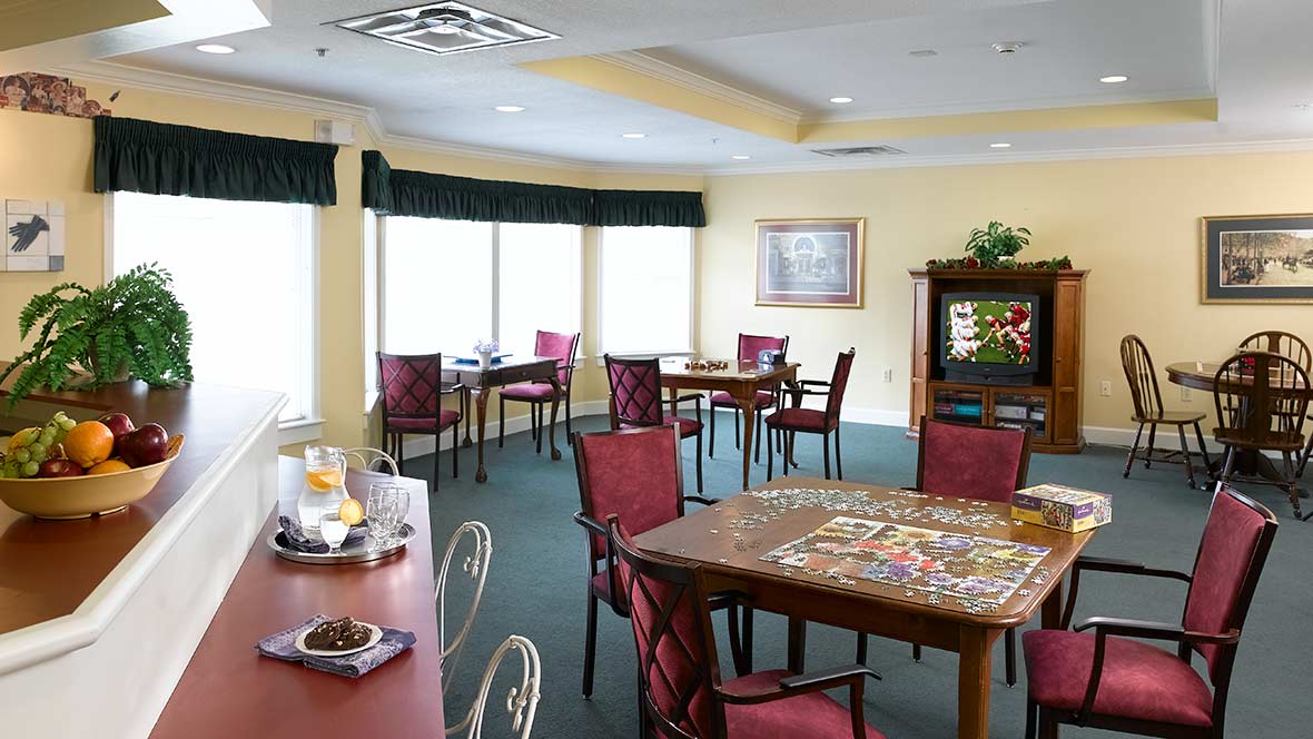 A cozy dining room with tables and chairs, featuring a puzzle on the table and large windows letting in natural light.