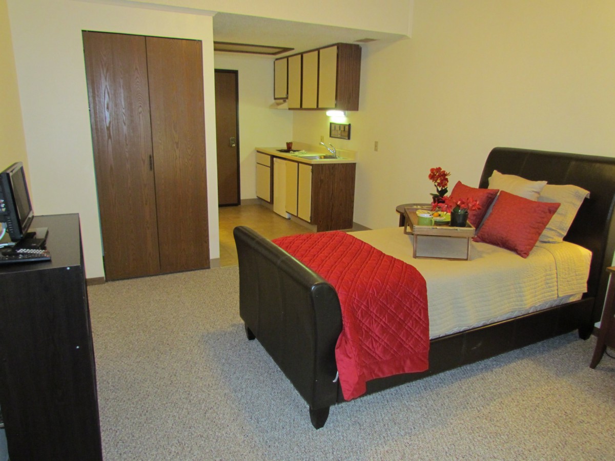 A cozy bedroom with a neatly made bed, adorned with red accents and a small kitchenette in the background.
