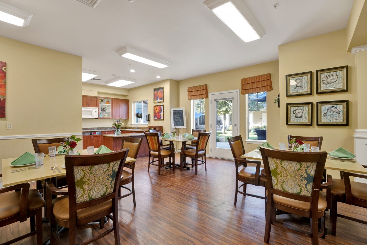 A cozy dining area with several tables and chairs, adjacent to a kitchen space, bathed in natural light from large windows.