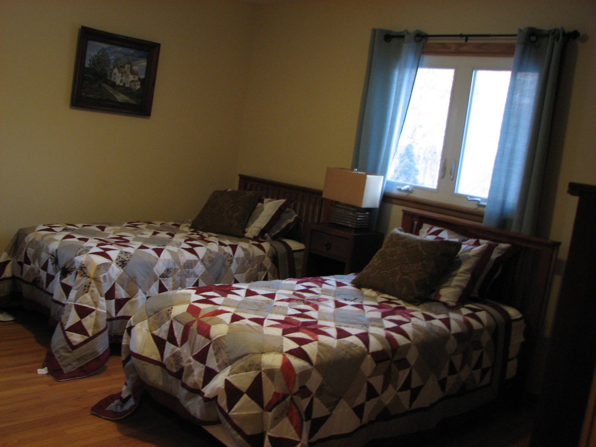 A cozy bedroom with two twin beds covered in patterned quilts, a window with blue curtains, and a small lamp on a nightstand.