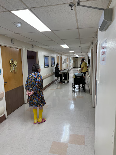 A scene in a hospital hallway where a woman stands while a caregiver assists a patient in a wheelchair.