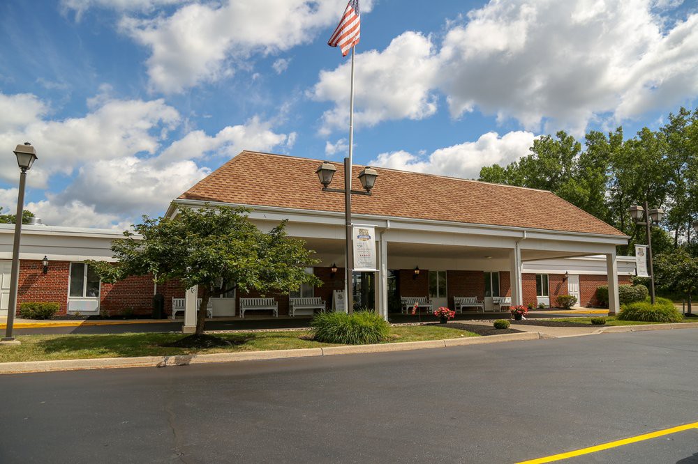 A front view of a hospital or clinic entrance with an American flag flying above, surrounded by trees and under a partly cloudy sky.