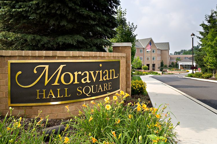 Entrance sign for Moravian Hall Square with a pathway and building in the background surrounded by flowers.