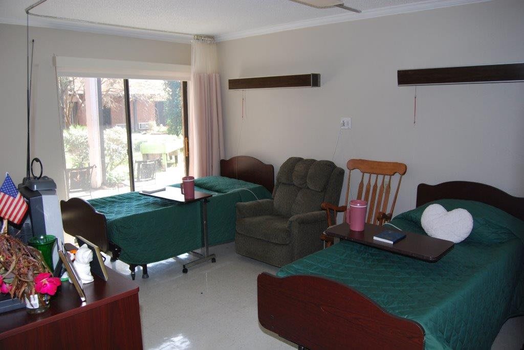 A shared nursing home or hospital room with two beds and seating area.