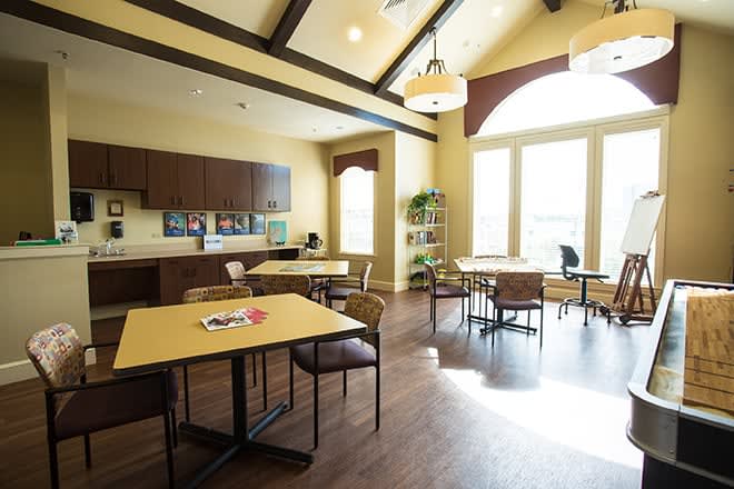 A well-lit dining area with tables and chairs, adjacent to a kitchen space with large windows allowing natural light.
