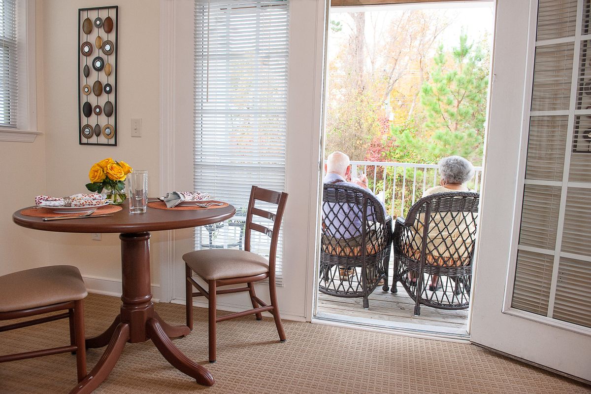 A cozy interior with a table and chairs leading to a balcony where two people are seated, enjoying the outdoor view.