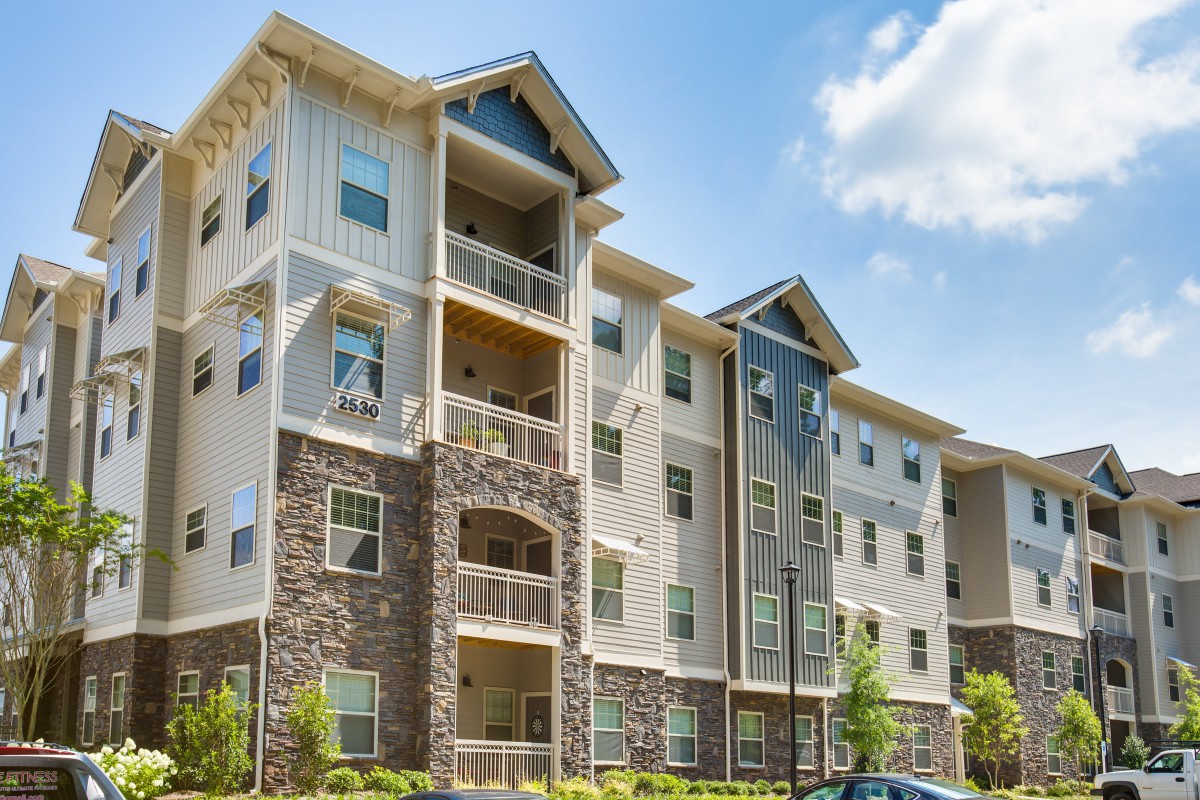 A modern multi-story apartment building with balconies and a stone facade, set against a partly cloudy sky.