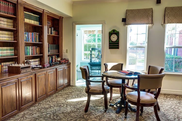 A cozy library room featuring a bookshelf, a round table with chairs, and sunlight streaming through the windows.