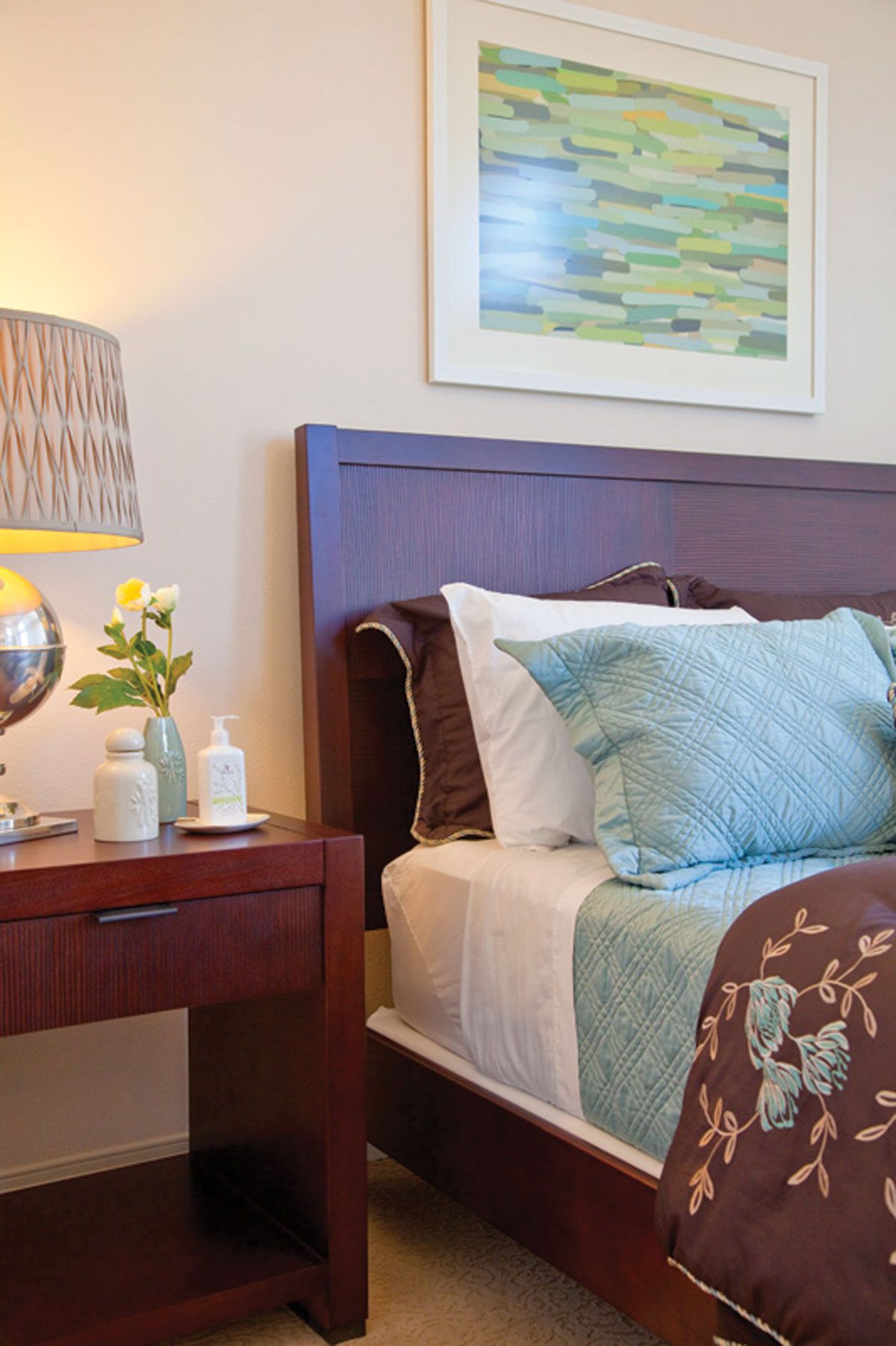 A cozy bedroom with a neatly made bed, decorative pillows, and a nightstand featuring a lamp and small plants.